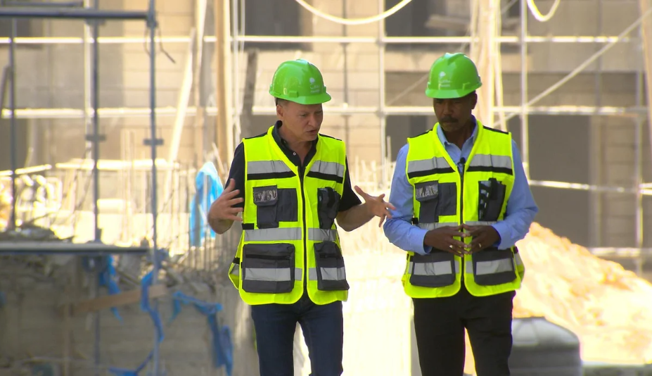Bashar Masri and 60 Minutes correspondent Bill Whitaker walking through a construction site in safety helmets and reflective vests